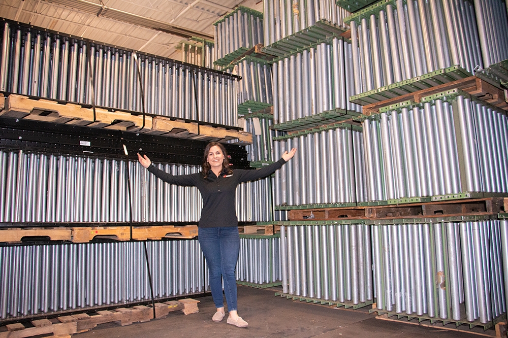 Woman standing in front of stacked used gravity conveyor rollers at a warehouse, showcasing inventory for sale