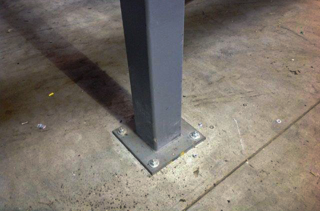 Picture of a Mezzanine Baseplate, sandwiched between a column and the concrete slab of a warehouse.