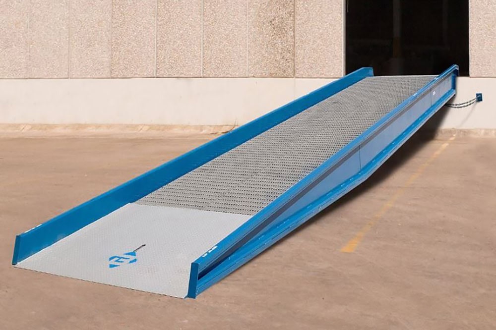 Blue dock-to-ground yard ramp connected to a warehouse loading dock, featuring non-slip metal decking and side safety curbs