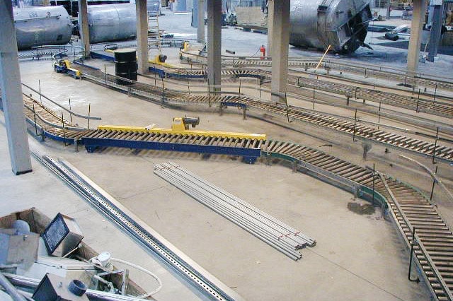 Industrial warehouse with a large gravity conveyor system, including multiple curved and straight roller conveyor sections arranged across the concrete floor for material handling and product transport.